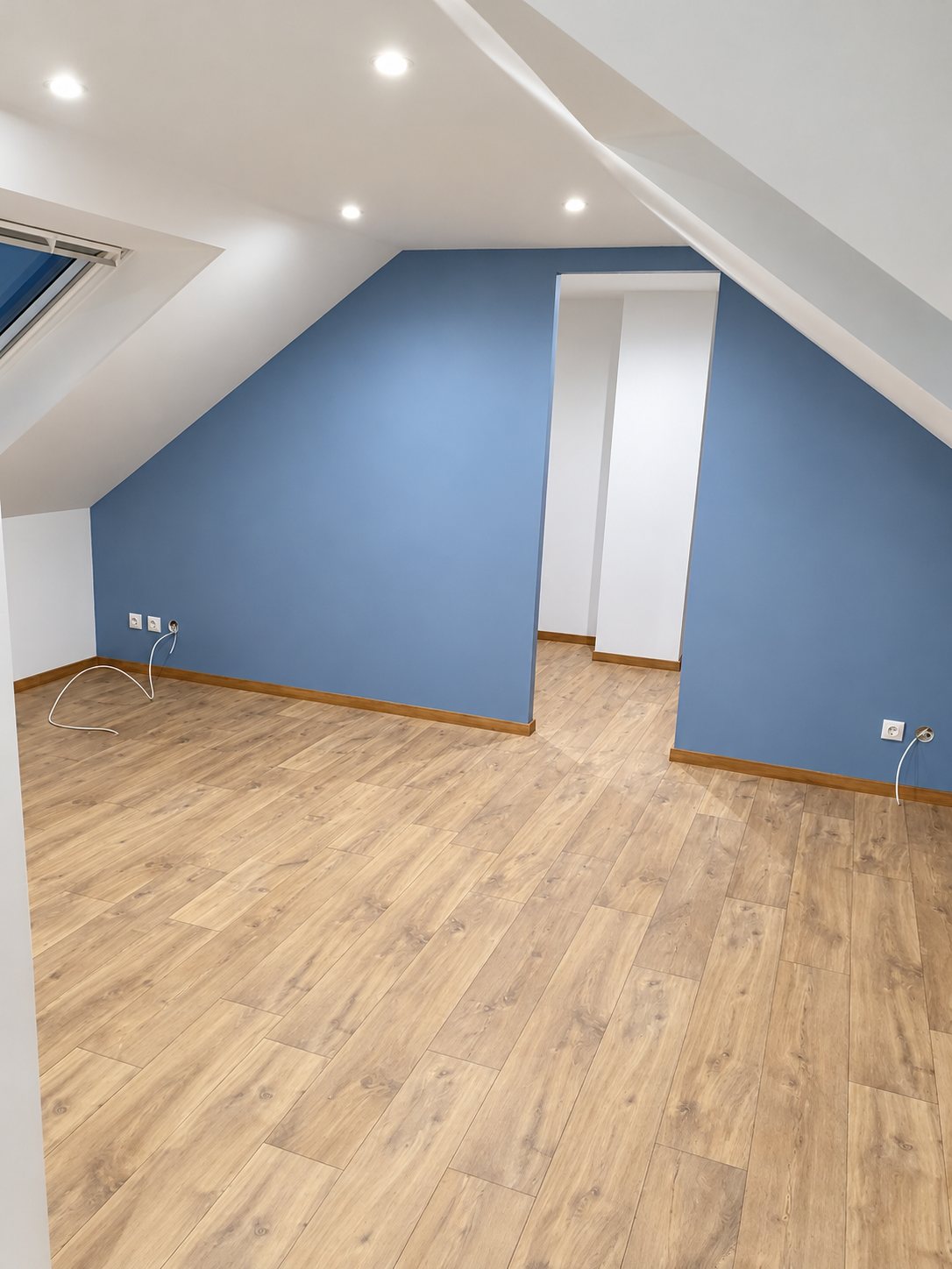 Pièce sous les combles rénovée avec murs peints en bleu, spots encastrés au plafond, plinthes en bois et sol stratifié clair.
