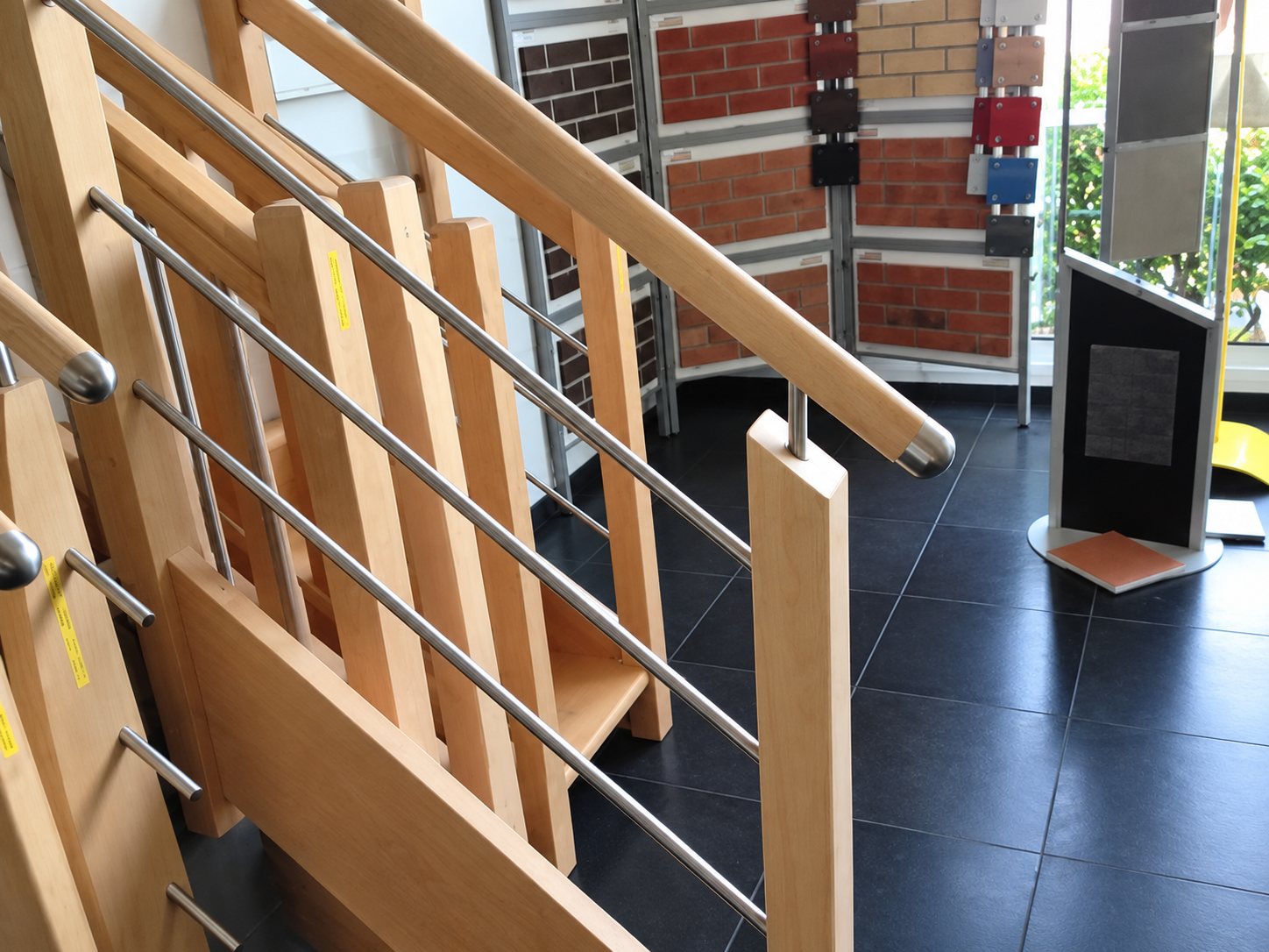 Un escalier en bois clair avec garde-corps à barres métalliques est exposé dans un showroom au sol sombre, avec des échantillons de matériaux au fond.