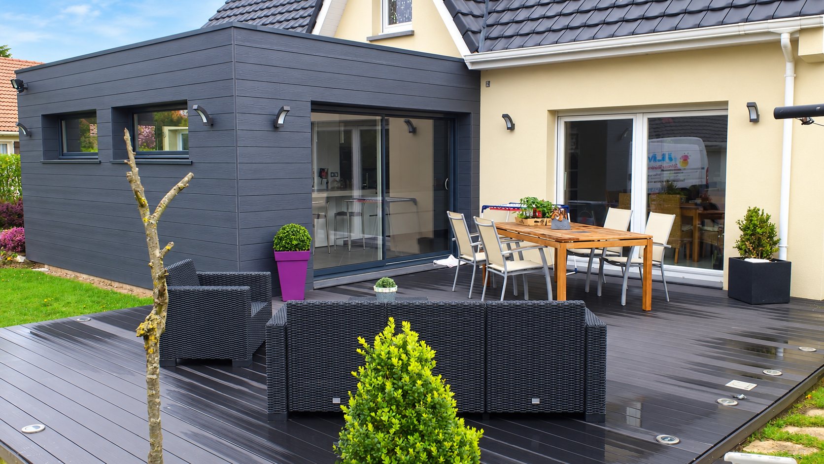 Une extension contemporaine à bardage gris anthracite s'ouvre sur une terrasse en bois composite meublée d'un salon de jardin en résine tressée et d'une table avec chaises.