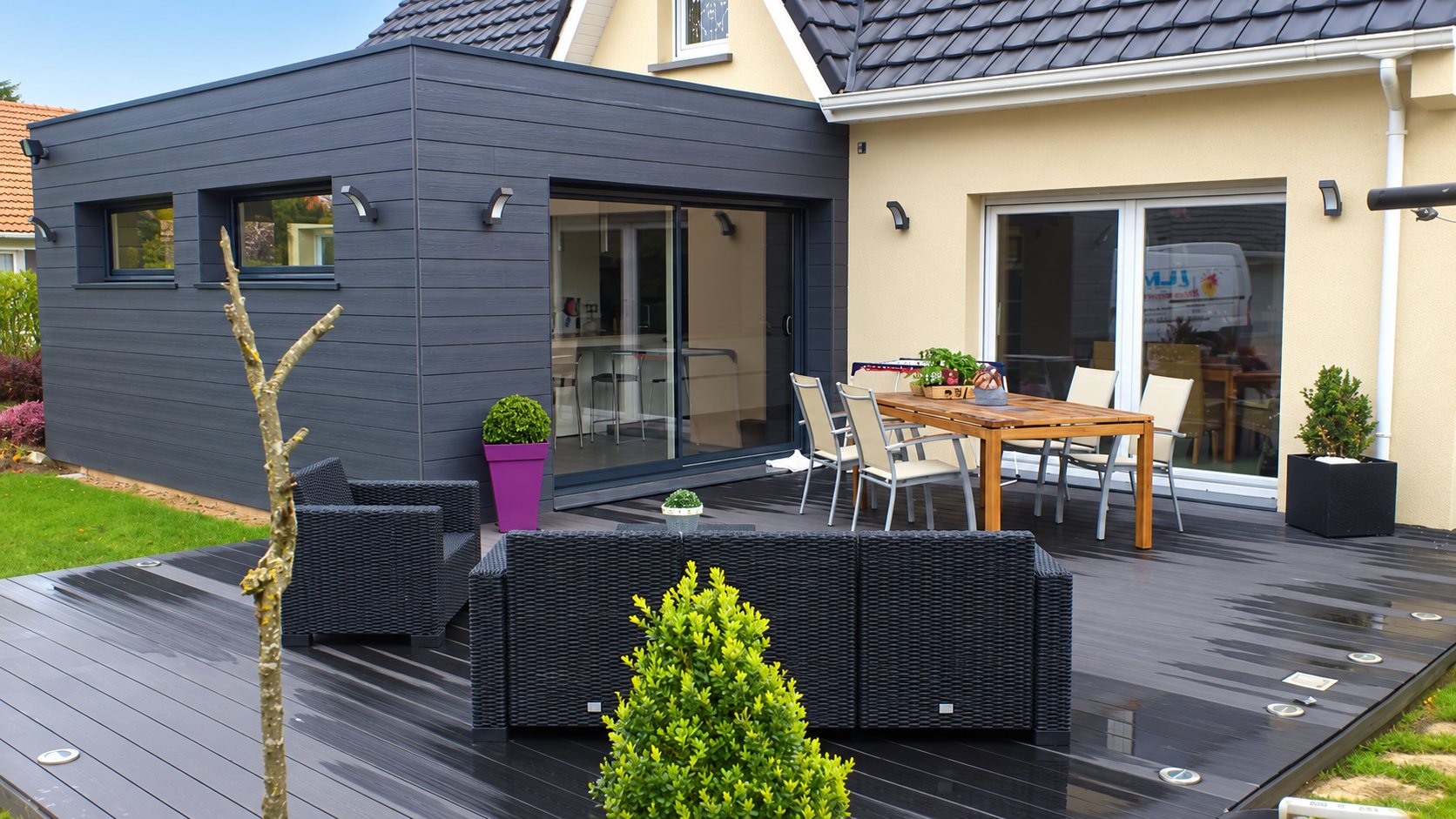 Une extension moderne au bardage gris anthracite s'ouvre sur une grande terrasse sombre meublée d'un salon en résine tressée et d'une table avec chaises, devant une maison crépie beige.