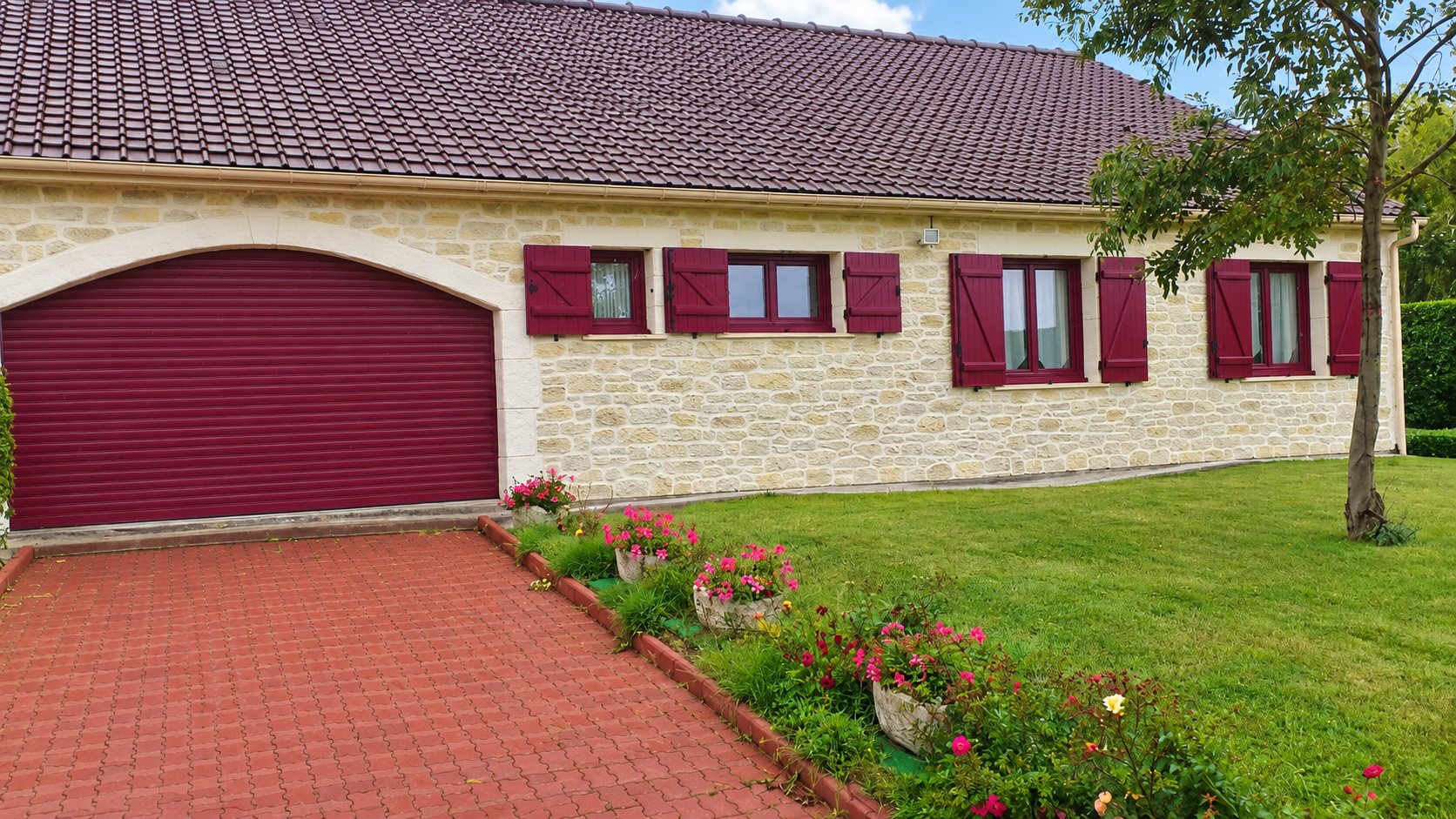 Façade d'une maison en pierre claire avec porte de garage roulante et volets battants bordeaux, devant une allée pavée rouge et un jardin fleuri.