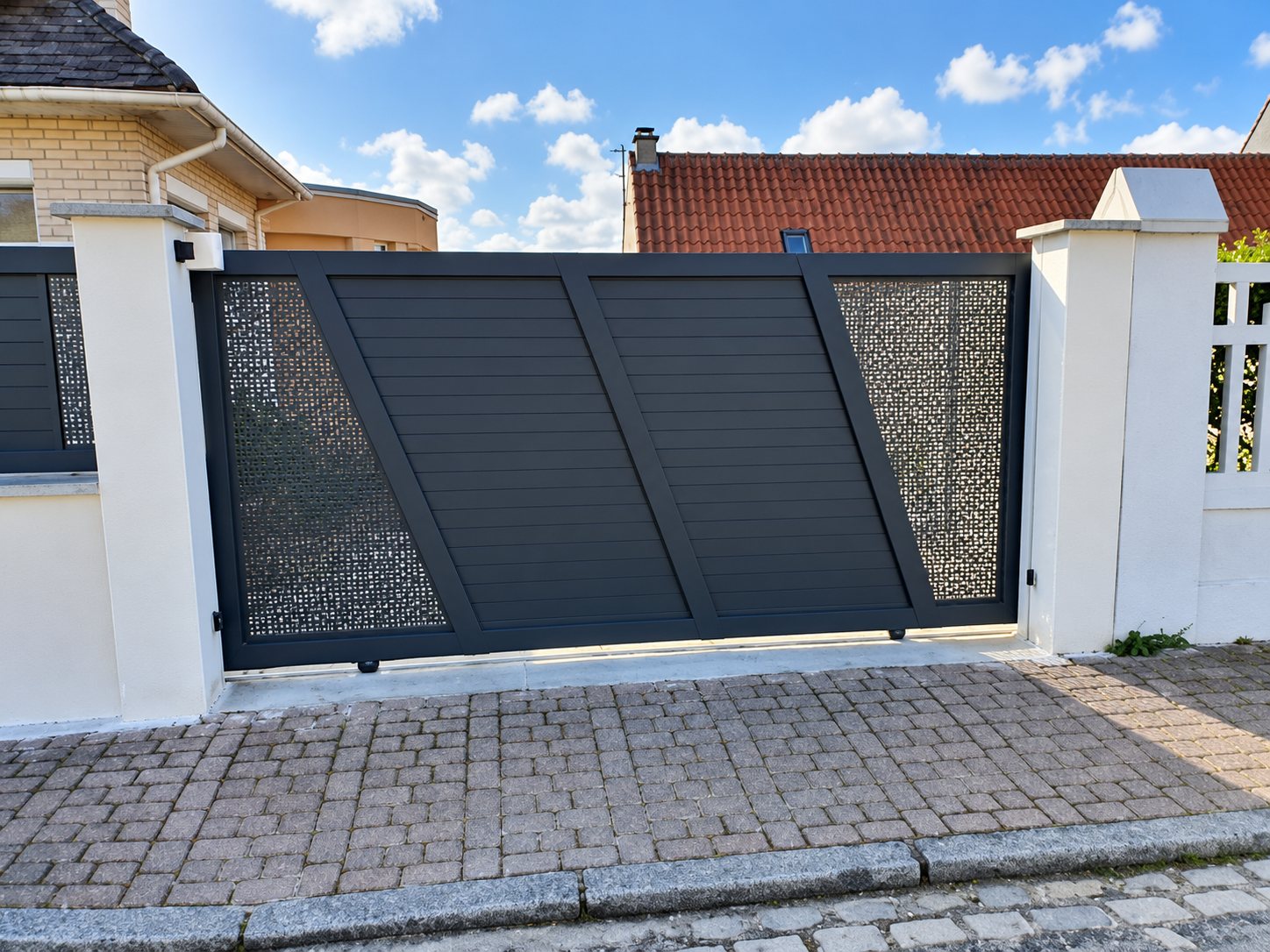 Un portail coulissant moderne gris anthracite à lames et panneaux perforés, encadré de piliers blancs, ouvre sur une allée pavée devant une maison.