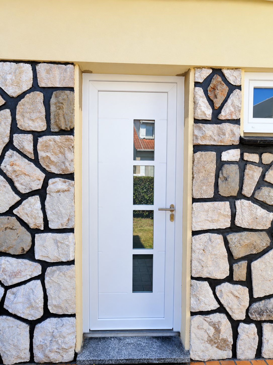 Une porte d'entrée blanche avec quatre petites vitres verticales et une poignée en inox, encadrée par un mur en pierres naturelles.