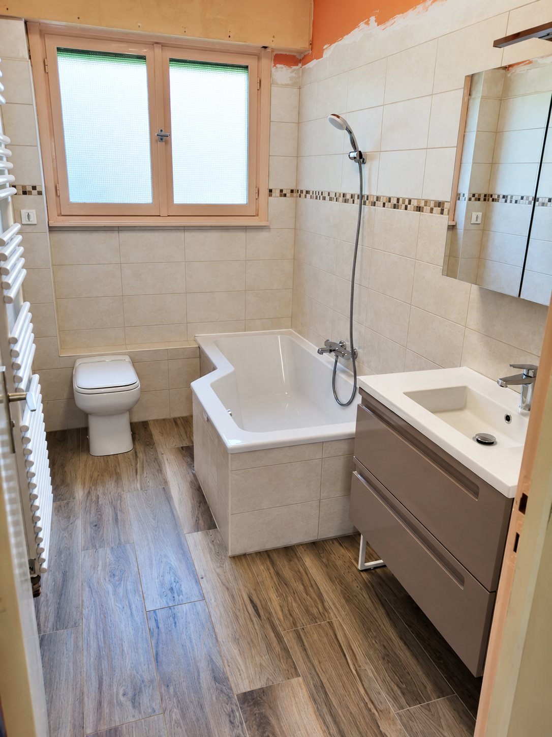 Salle de bain récemment rénovée avec baignoire d'angle, meuble vasque suspendu, WC, sèche-serviettes et sol imitation bois.
