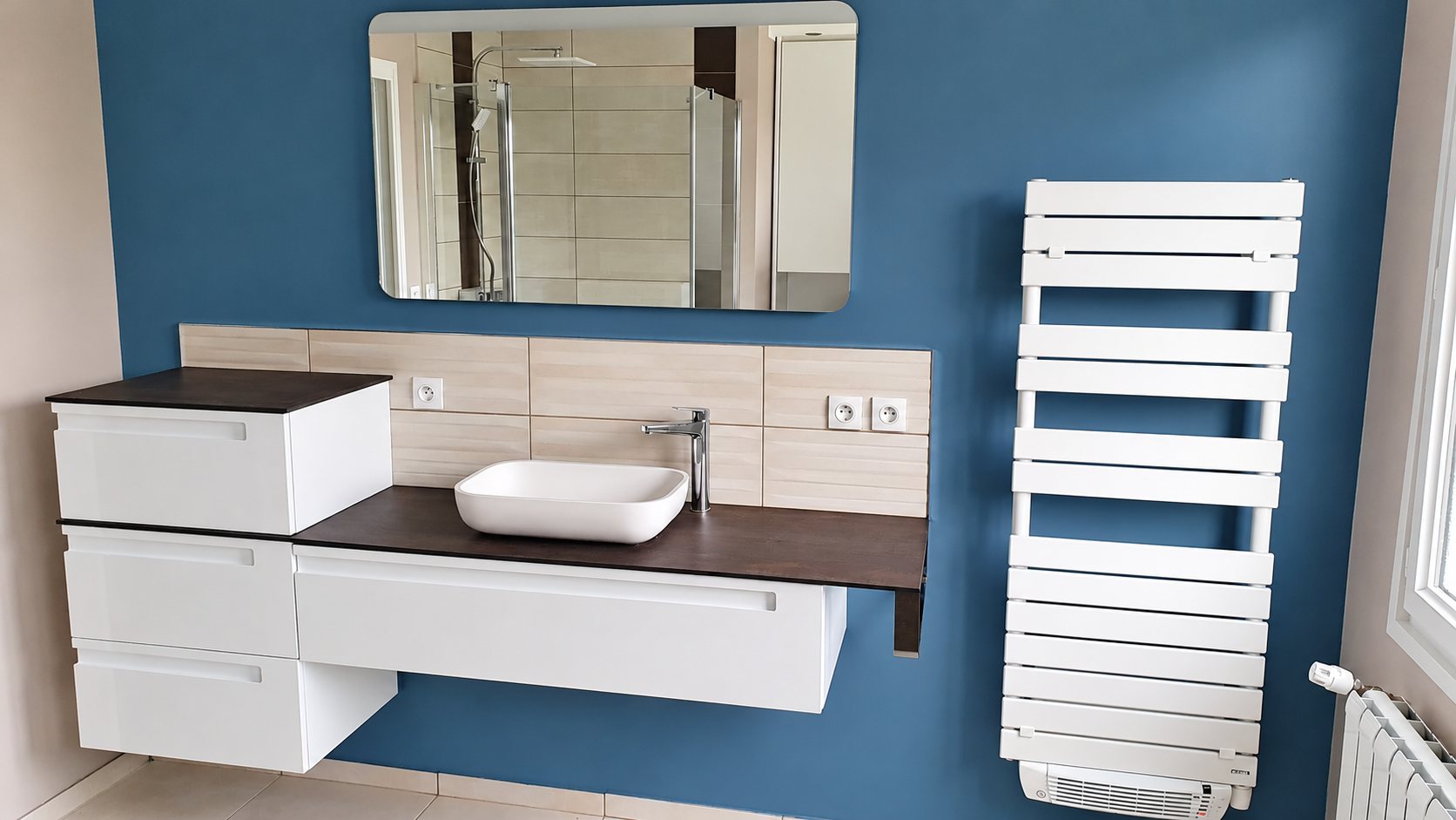 Salle de bain aux murs bleu-gris avec meuble suspendu blanc, vasque à poser, grand miroir et sèche-serviettes blanc.